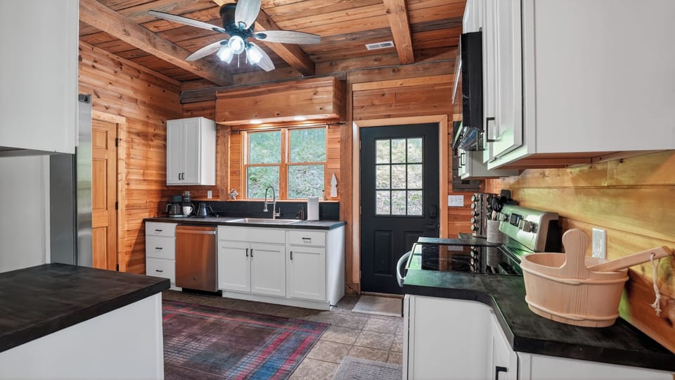 White shaker cabinets, dark countertops, stainless appliances, a window into the treeline, and a back door that opens to the deck. A fully stocked kitchen that actually means it.