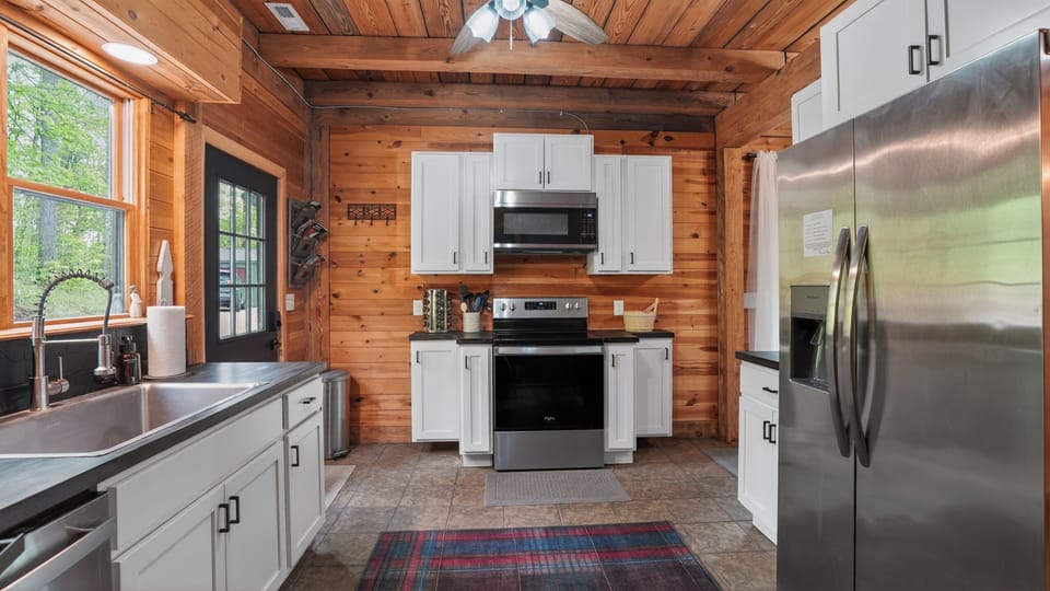 Full stainless suite — fridge, range, microwave, dishwasher. White cabinets, dark counters, plaid rug underfoot. Mornings here smell like coffee and cedar.  