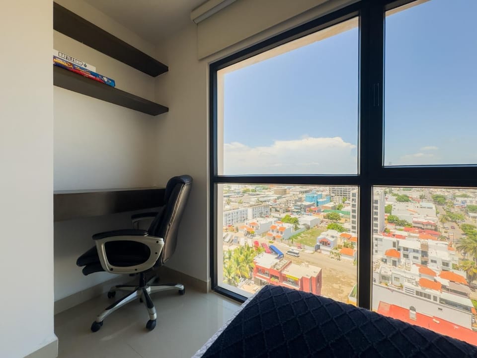Small office nook with dark wood desk and shelves, black swivel chair, and large window overlooking city rooftops.
