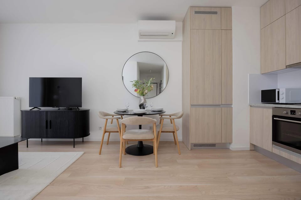 An open layout displays a dining section with a round black table beneath a circular mirror. Behind the seating area, seamless wooden cabinetry houses built-in kitchen appliances. Light wood floors tie the space together.