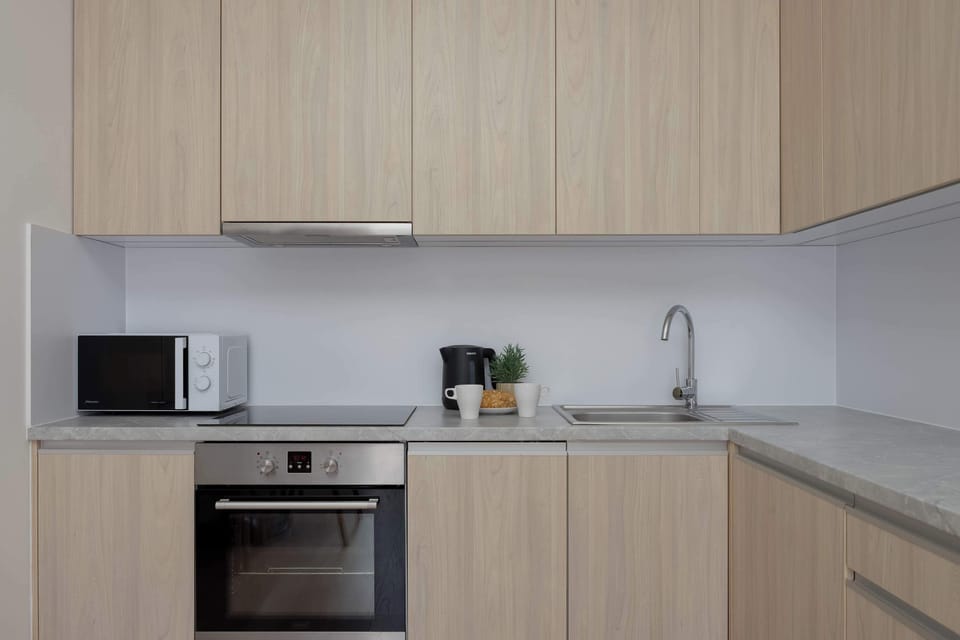 A clean kitchen counter setup features light wooden cabinetry and built-in appliances. A sleek sink sits beside a modern cooktop and microwave. Light grey countertops create a clean, contemporary aesthetic.