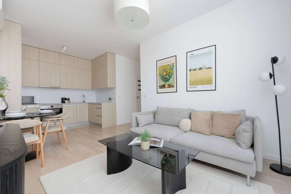 A spacious living room combines a plush grey couch with a modern black coffee table. Two framed botanical prints decorate the far wall above the dining zone. The open layout connects seamlessly to a light-wood kitchen.