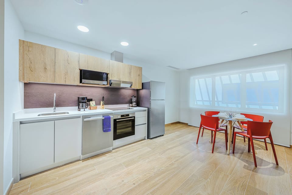 Your fully equipped kitchen and dining area features modern appliances and a bright, open layout enhanced by expansive floor-to-ceiling windows. Bright red dining chairs add a vibrant splash of color, creating a warm and inviting space to enjoy every meal.