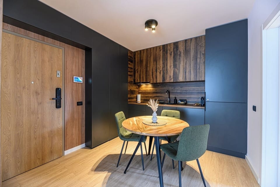 Open-plan kitchen and dining area with anthracite cabinetry, full-height rustic wood panelling, round dining table and 3 upholstered green chairs. Smart lock entrance door visible alongside. Grand Chalet Suite 2, Poiana Brasov.