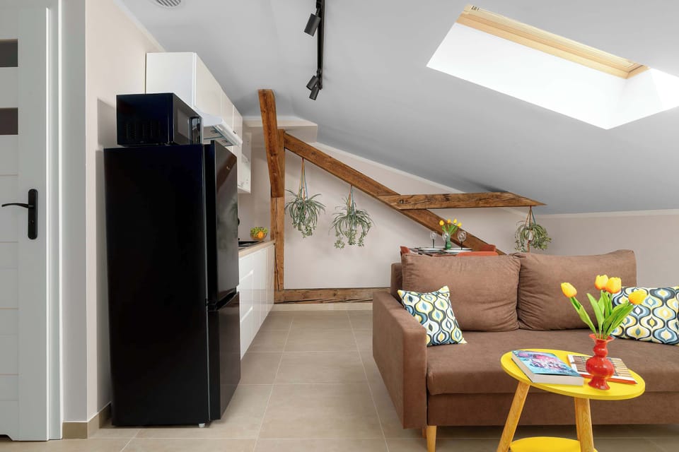 An entry view of the apartment shows a black refrigerator on the left and a brown sofa visible under the exposed wooden beams of the ceiling. A yellow side table brings a cheerful accent to the space.