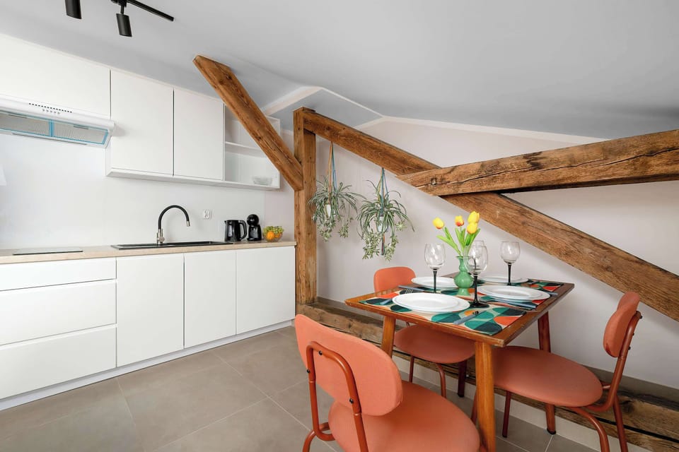 A dining space features a wooden table with orange chairs, positioned next to a white kitchen counter under exposed wooden beams. Hanging plants add a touch of nature to the wooden framework.