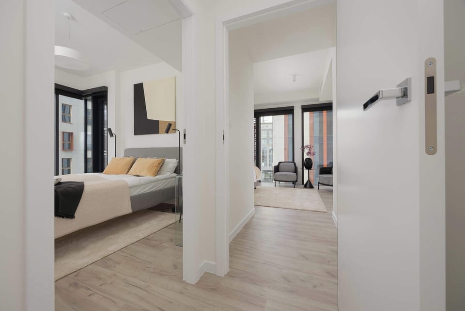 A bright hallway with light wooden flooring leads directly into a cozy bedroom. The white doors and neutral walls give the entrance a clean and welcoming appearance.