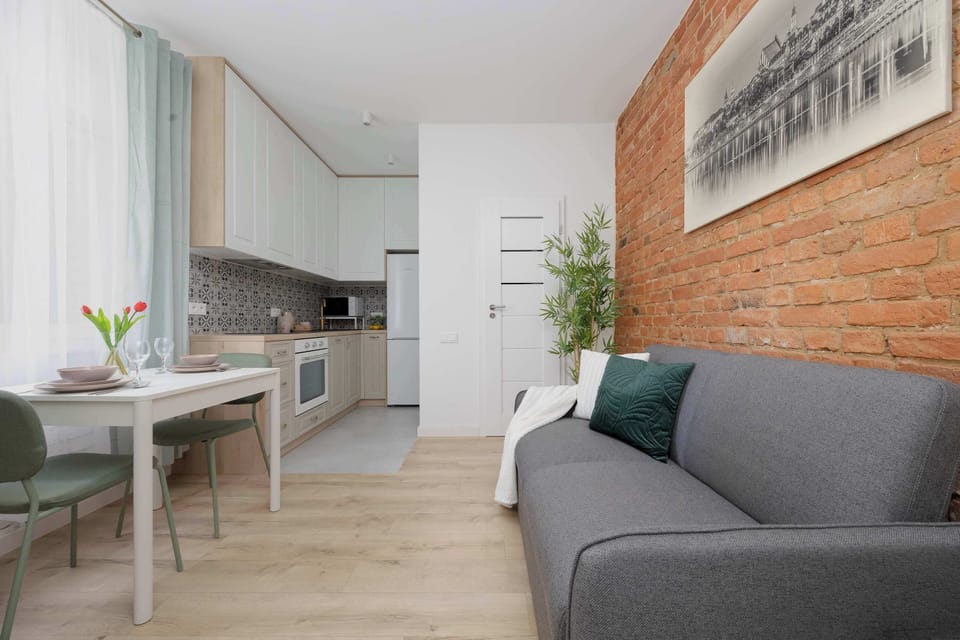 An open-plan space includes a kitchenette, dining area, and a gray sofa. Light flooring and brick walls give it a modern look.