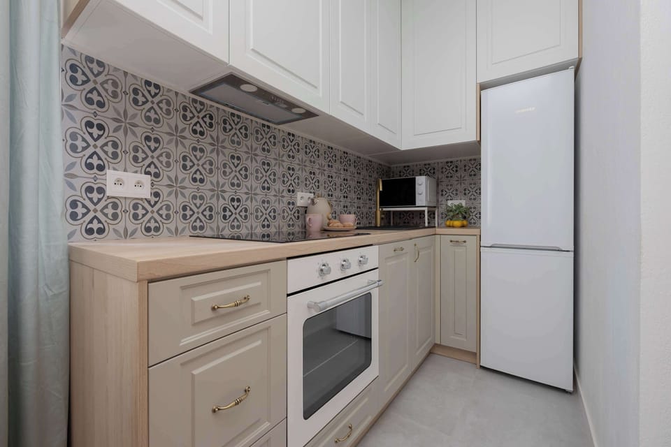 A modern kitchenette includes white cabinets, an oven, and a patterned backsplash. A tall refrigerator stands at the end of the counter.