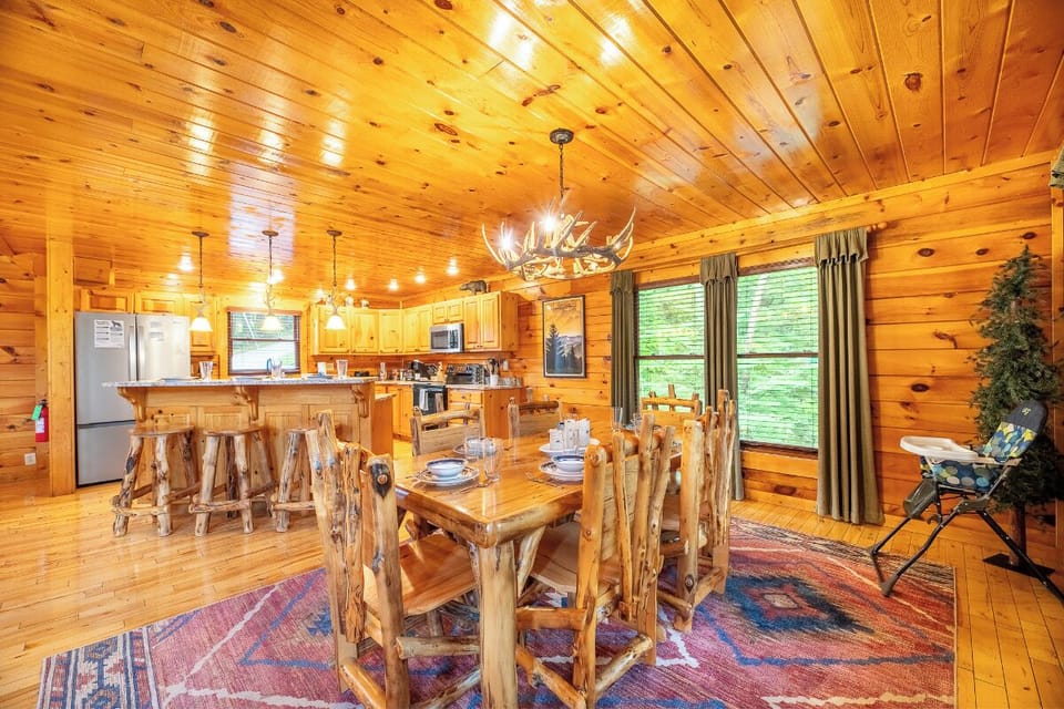 Gather around rustic log furniture in your open-concept cabin, where warm wood surrounds you and natural light streams through large windows overlooking peaceful greenery.