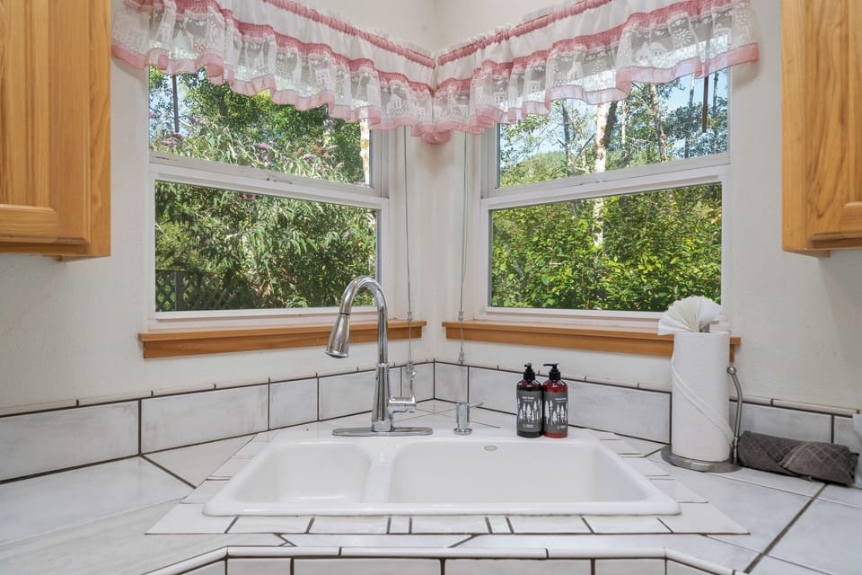 A bright kitchen corner sink framed by sunlit windows overlooking lush greenery, with handsome tile countertops and warm wood cabinetry creating a cozy, cottage-style feel.
