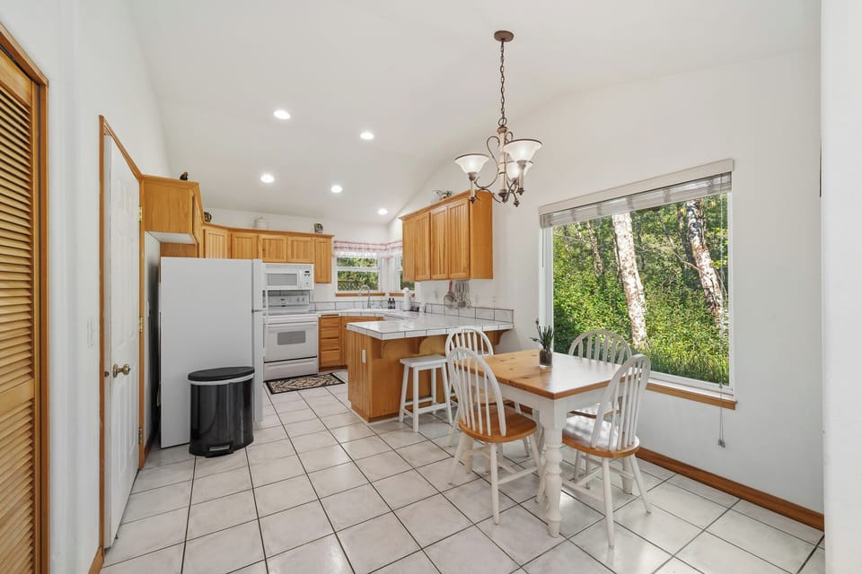 Gather around the dining table in this sunny kitchen where natural light streams through windows overlooking lush greenery. The fully equipped space invites you to prepare meals and share stories.