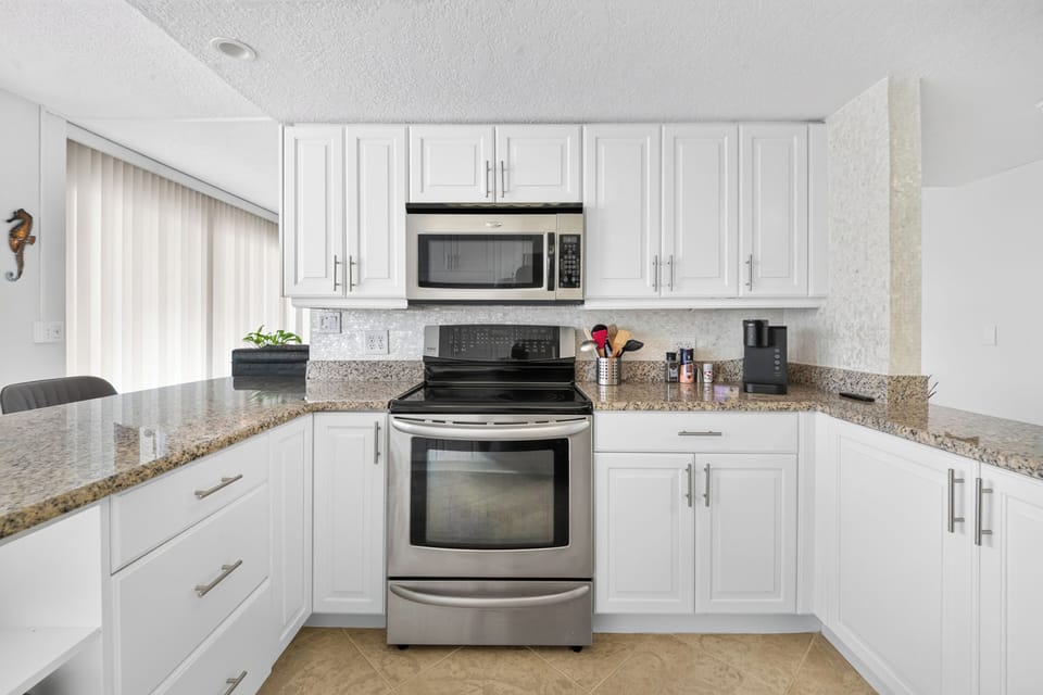 Your fully equipped kitchen features sleek white cabinetry, granite countertops, and modern stainless steel appliances—everything you need to prepare meals or brew morning coffee in comfort.