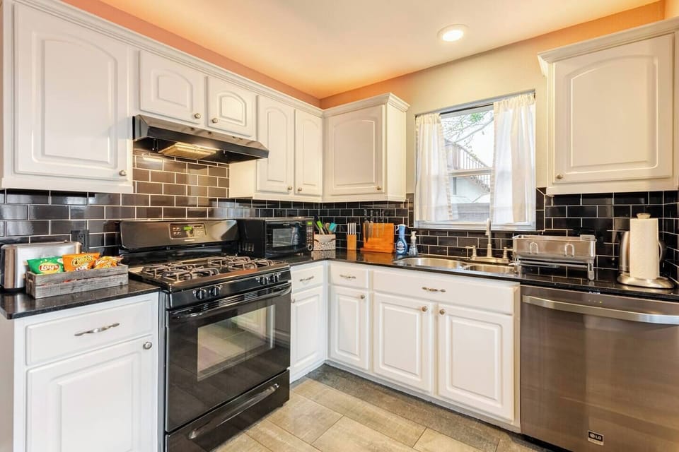 Fully equipped kitchen with black appliances, gas stove, microwave, and dishwasher. Black tile backsplash complements white cabinets, and a sunny window adds charm. Perfect for cooking meals or enjoying provided snacks during your stay.