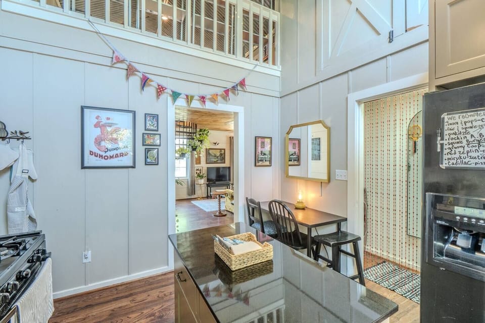 A dining nook with a slim black table and two chairs against a light grey wall with a gold mirror. High ceilings feature a loft overlook and decorative bunting. A black granite kitchen island and a black refrigerator frame the space.