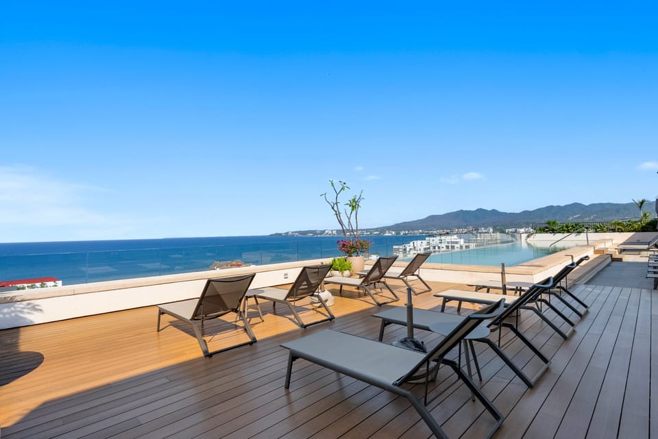 Panoramic rooftop sun deck featuring sleek lounge chairs, an infinity pool, clear glass railings, and unobstructed views over the ocean and mountains.
