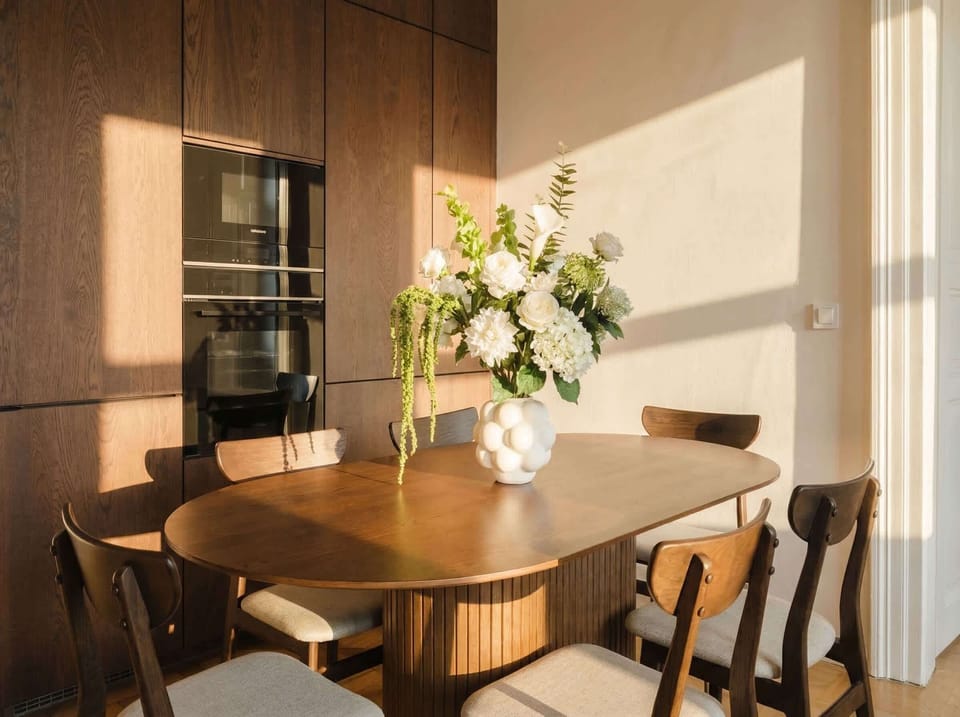 Sunny dining area with a wooden table and a fully equipped kitchen nearby. A pleasant space for breakfast, coffee, or a relaxed meal at home.