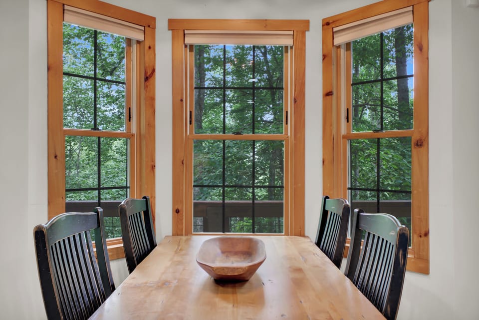 Three walls of windows turn this dining nook into a treehouse — every pane framing solid green canopy in summer, and come October, that same view ignites into gold, orange, and red without you leaving your seat.