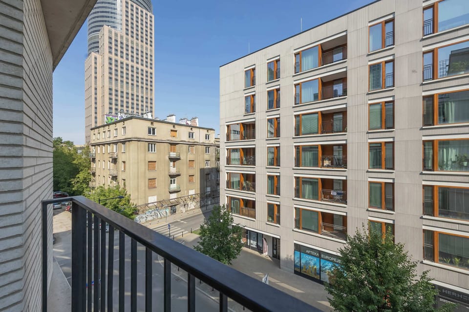 A high-angle balcony view looks across at neighboring modern apartment facades and a tall city building under a bright blue sky.