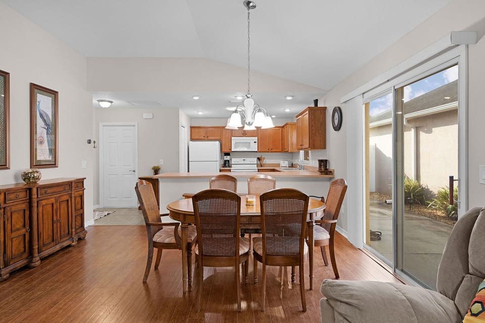 Elegant dining table beneath vaulted ceilings and soft chandelier light, with an adjacent kitchen and seamless flow to the living space.