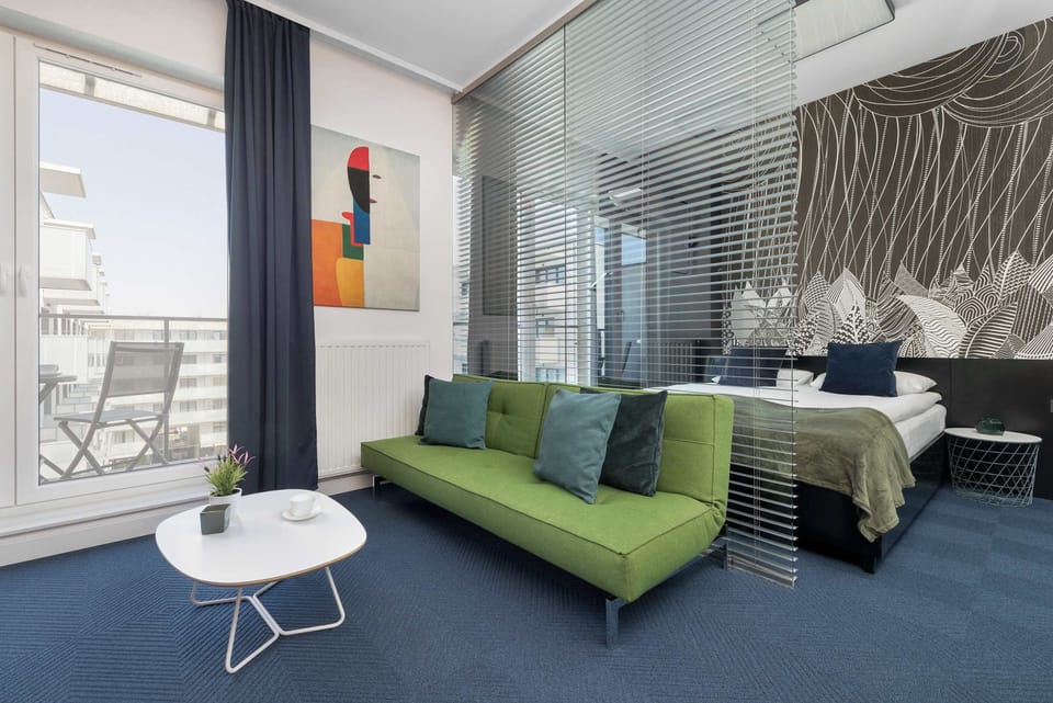 A wide shot of an open-plan living space combining a lounge area, dining space, and a sleeping corner. A vibrant green sofa and a white dining set sit on a blue carpet.