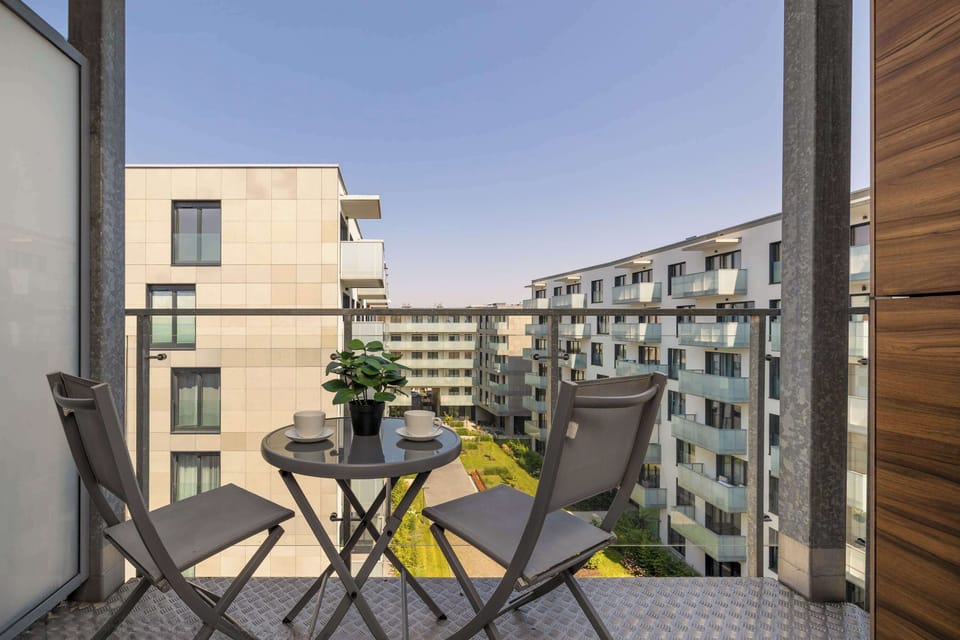 A balcony equipped with a small, round table and two folding chairs for outdoor relaxation. The view overlooks a modern apartment building complex under a clear sky.