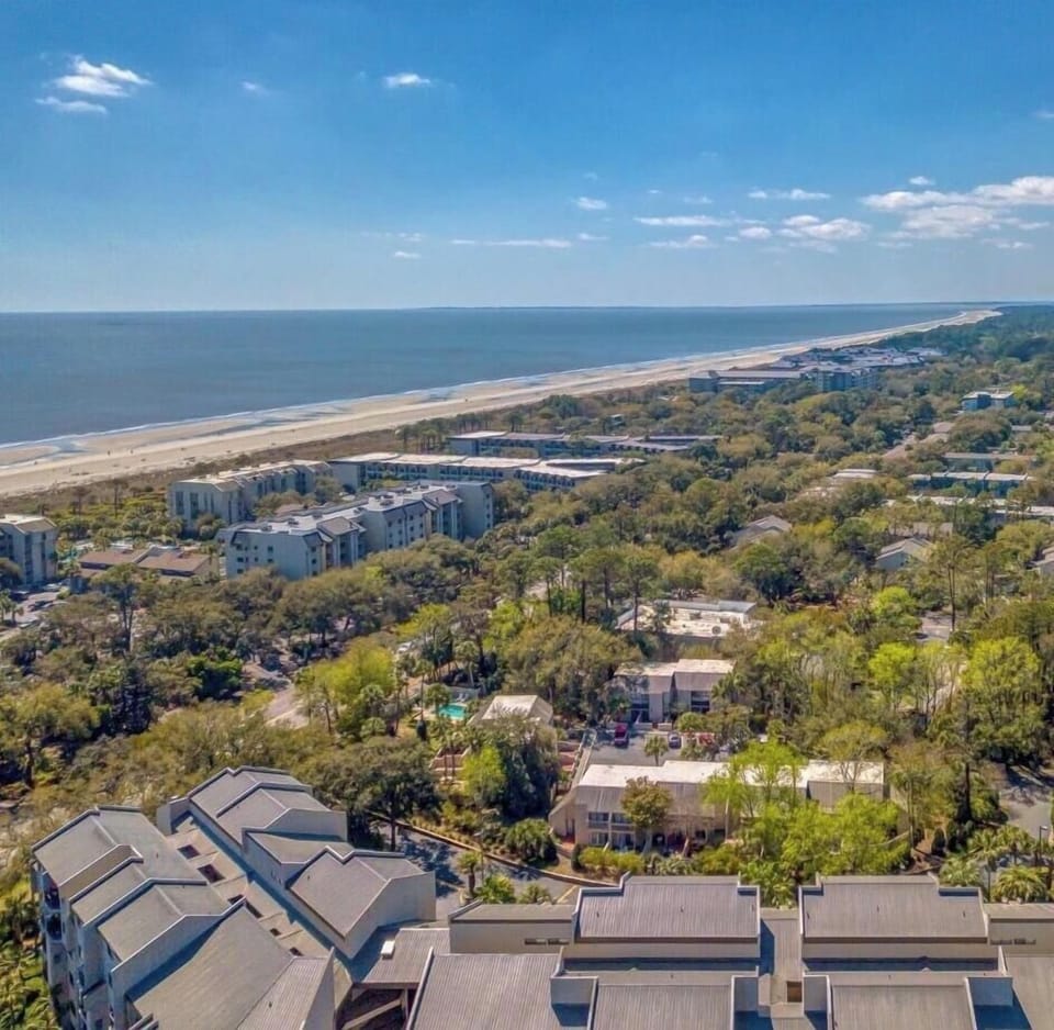 Vibrant greenery meets the deep blue horizon in this expansive aerial view. The saltwater breeze drifts over a dense canopy of trees and quiet rooftops, promising a peaceful stay just moments away from the rolling surf and wide sandy shores.