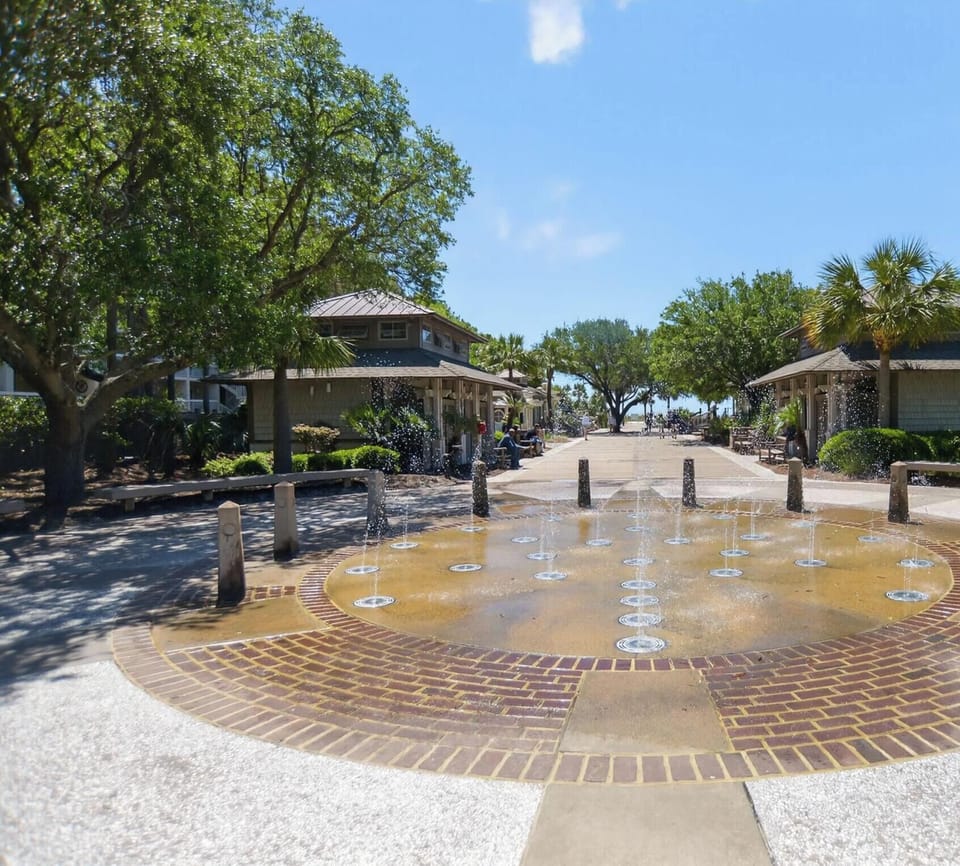 Sparkling water jets dance above the warm brickwork of this sunny plaza, creating a playful path toward the beach. Shady trees and classic coastal architecture frame a serene walk under the wide, blue summer sky.