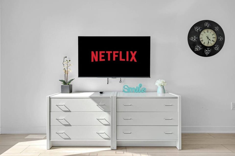 A white media console table underneath a wall-mounted television displaying Netflix, flanked by a decorative plant and clock—ideal for setting out snacks and hosting a fun movie night.