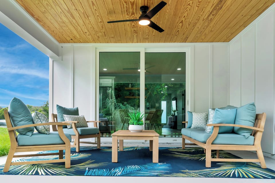 Step outside to serene patio living! 🌿 Relax in cushioned comfort under the breezy ceiling fan while enjoying surrounding greenery and coastal decor. 🍹☀️