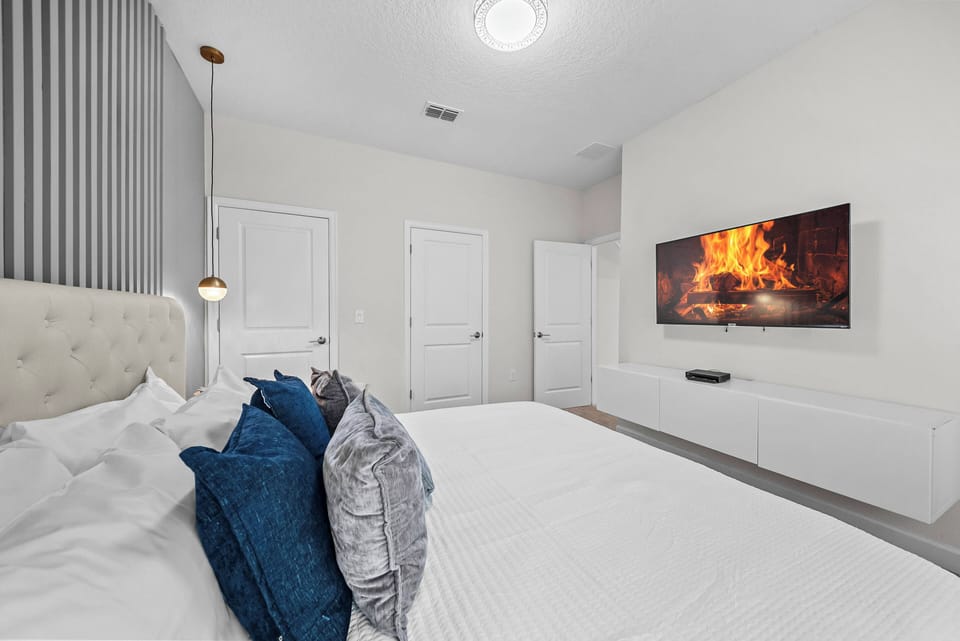 Welcome to a serene bedroom featuring soft neutral tones and plush bedding with rich blue pillows. Enjoy entertainment on a sleek wall-mounted TV, and bask in the natural light that fills this inviting and cozy retreat.