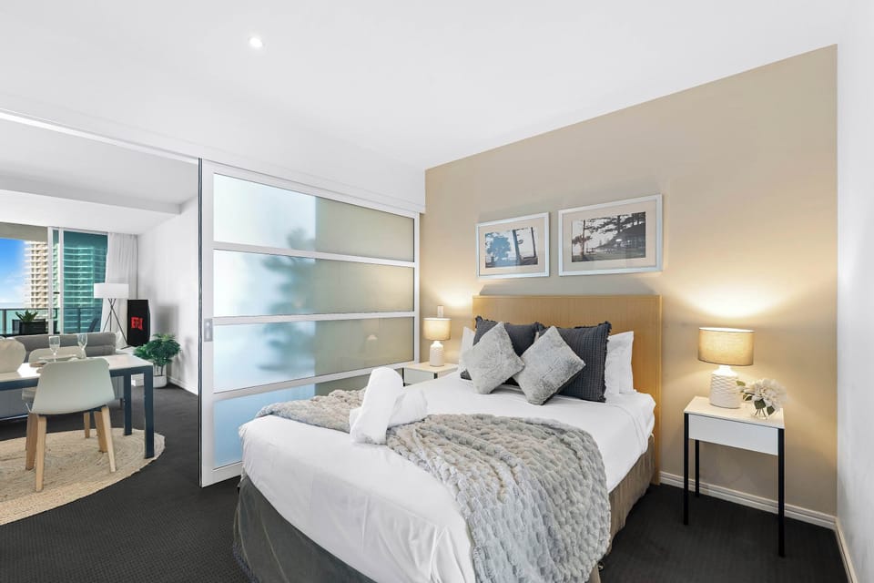 Rest easy in this welcoming bedroom space, featuring a comfortable bed flanked by modern nightstands, framed art overhead, and frosted glass sliding partitions that provide separation from the adjoining dining and living area.