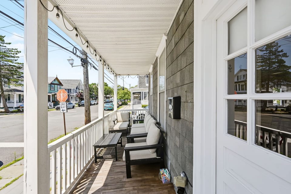 Enjoy your morning coffee or relax in the evening on this covered front porch, featuring comfortable outdoor seating with plush cushions, festive string lighting, and views of a quiet, walkable neighborhood street.