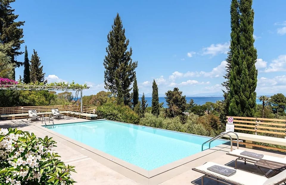 A sunlit outdoor swimming pool features lounge chairs and tall cypress trees overlooking a scenic sea view.
