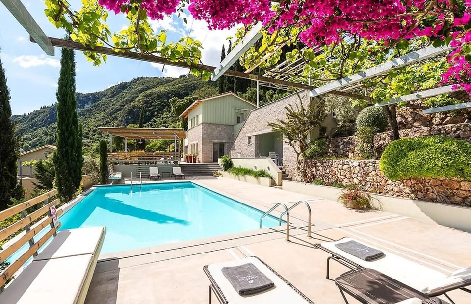Bright pink bougainvillea frames a view of the outdoor swimming pool, terrace lounge chairs, and the multi-level stone villa built into a green hillside.
