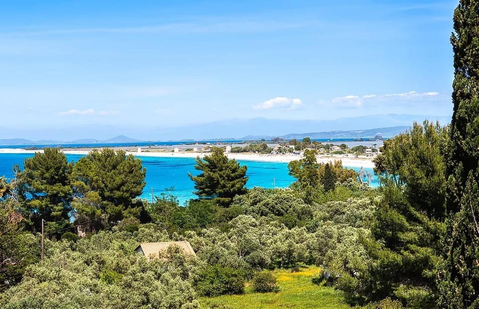 A panoramic viewpoint highlights a sandy beach and turquoise sea, framed by a dense canopy of green trees under a clear blue sky.