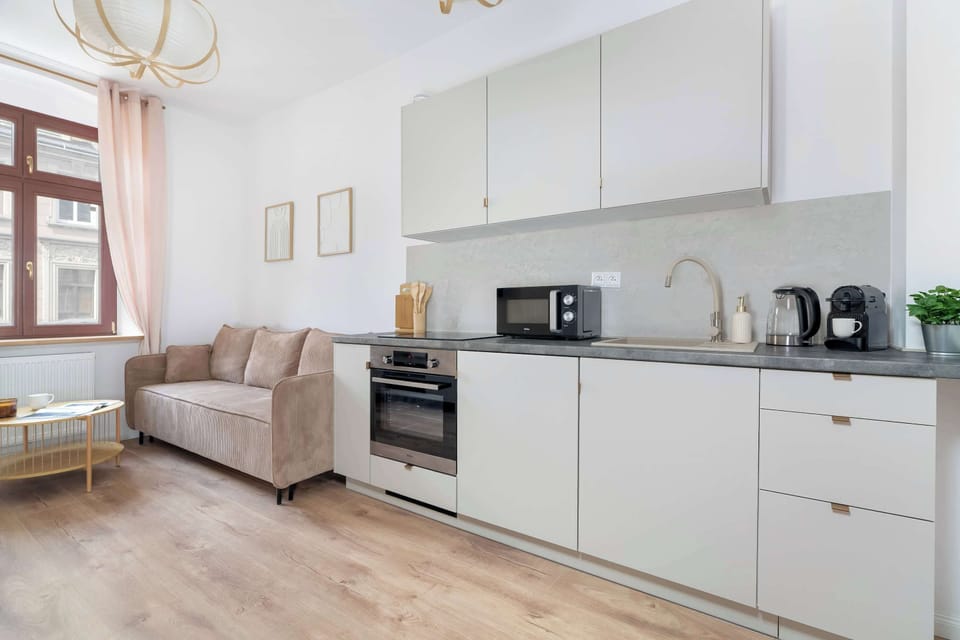A linear kitchenette is equipped with light grey cabinets, a black microwave, and an integrated oven. A beige sofa sits in the open living space opposite.