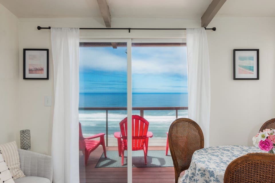 Step through glass doors onto your private deck where vibrant red chairs frame an endless ocean horizon—your morning coffee spot with the sound of waves.