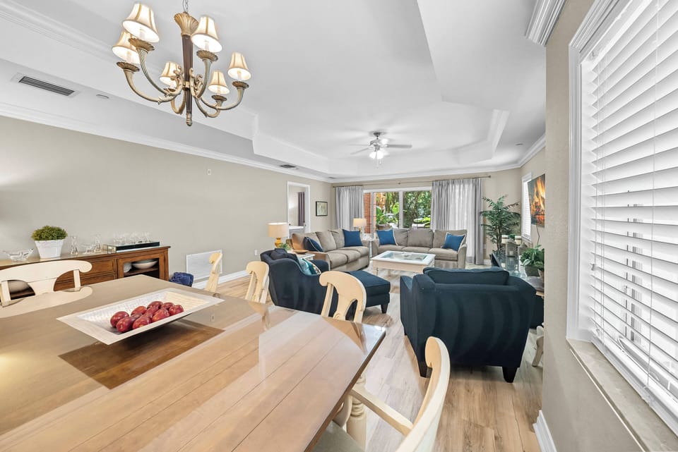 Enjoy a bright, airy living area with warm wood tones and cozy deep blue sofas. A stylish chandelier enhances the ambiance, while large windows invite natural light and views of the lush outdoors, perfect for relaxing gatherings.