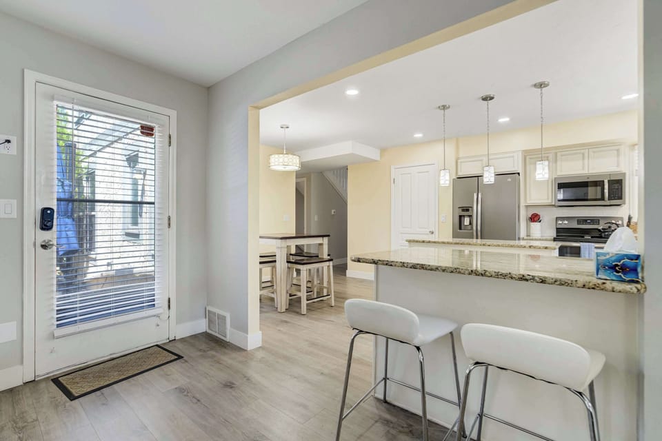The kitchen opens off the living area behind a breakfast bar set with modern stools and lit by a row of clear glass pendants. A keypad entry door leads straight out to the patio, keeping the grill within easy reach of the counter.