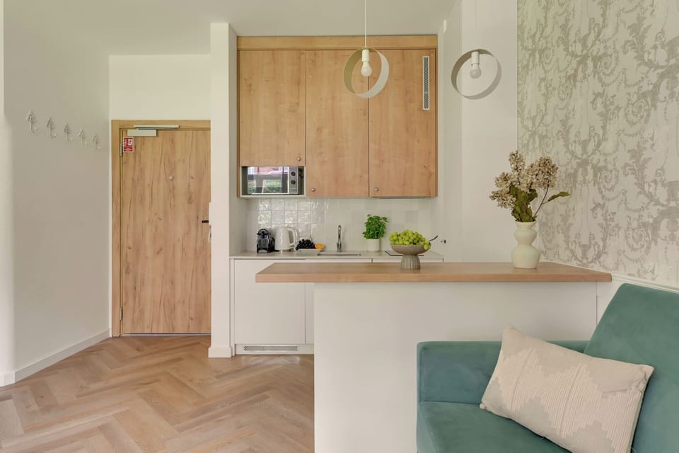 Jasny i przytulny salon z wygodną sofą, dużymi oknami i elegancką podłogą ułożoną w jodełkę. Delikatne dekoracje i stonowana kolorystyka tworzą harmonijną, komfortową przestrzeń do wypoczynku.