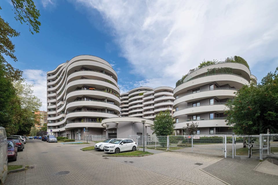 An exterior view of a modern residential complex with curved balconies and landscaped surroundings. Several cars are parked near the entrance of the building.