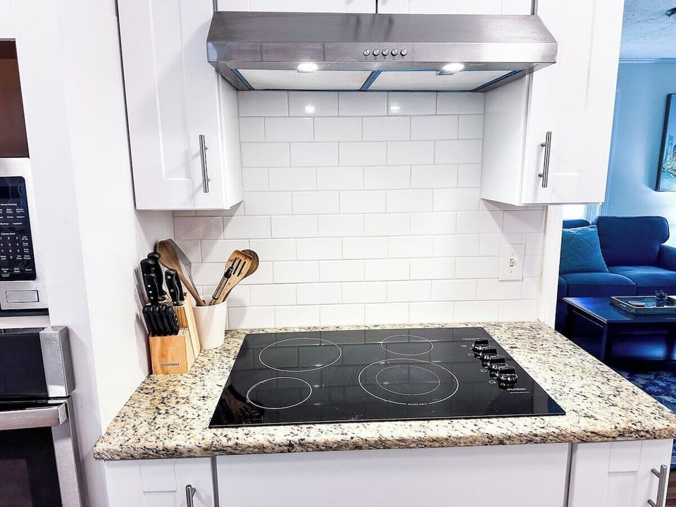 Cook up delicious meals using this sleek electric cooktop, set into granite countertops with white subway tile backsplash, white cabinetry, and a stainless steel range hood.