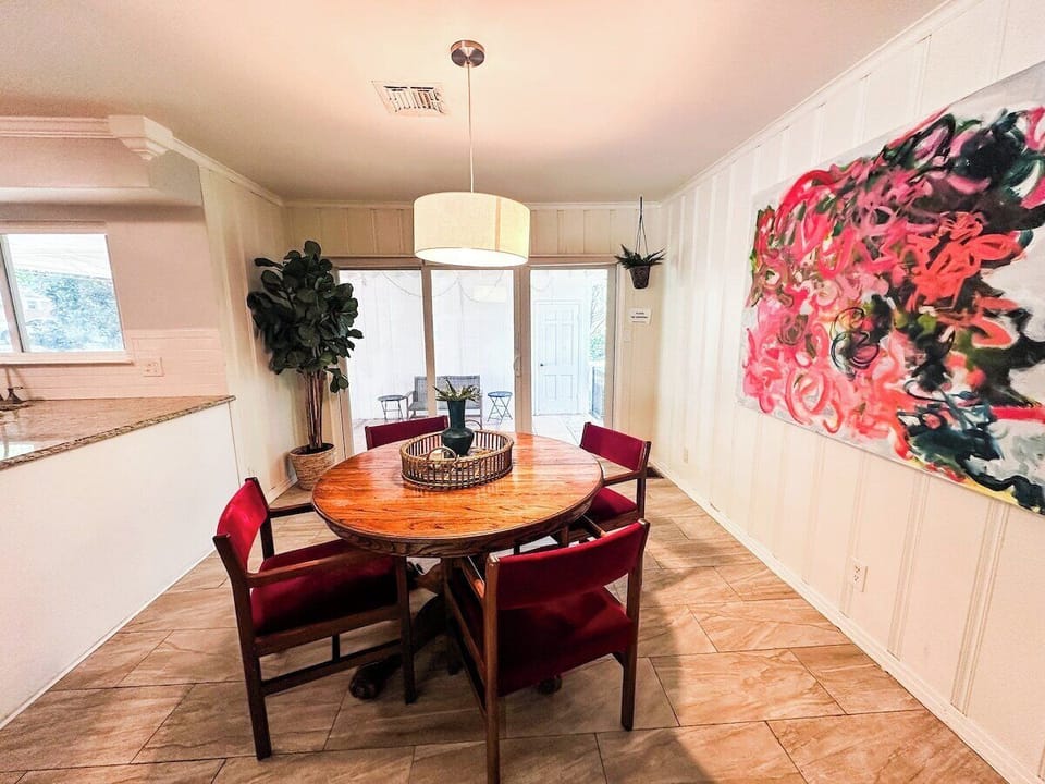 Gather for meals in this bright dining space, featuring a round wooden dining table paired with red upholstered armchairs, a modern drum pendant light, vibrant large-scale abstract artwork, and sliding glass doors leading to the patio.