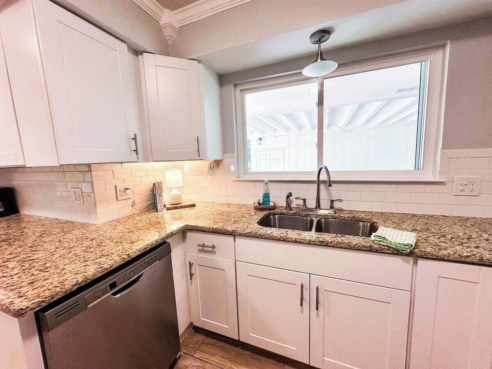 Enjoy cooking in this bright kitchen corner, featuring crisp white shaker cabinets, granite countertops, a stainless steel dishwasher, an undermount double sink set below a wide window, and a subway tile backsplash.