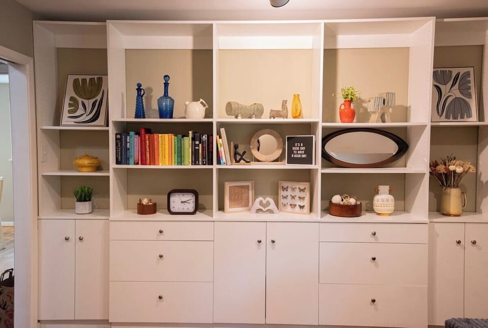 Store and display items in this large white built-in wall unit featuring open shelves decorated with books, framed art, and decorative accents, along with lower cabinets and drawers for additional storage.