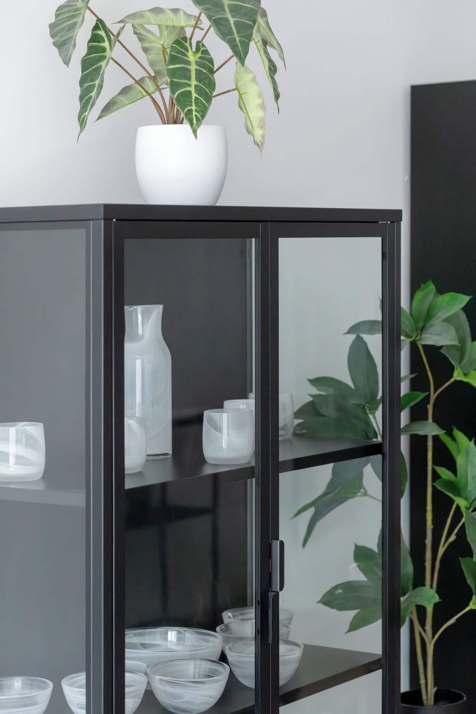 Close-up view of a sleek dark display cabinet with glass doors. Decorative white tableware and green house plants rest on the glass shelving inside.