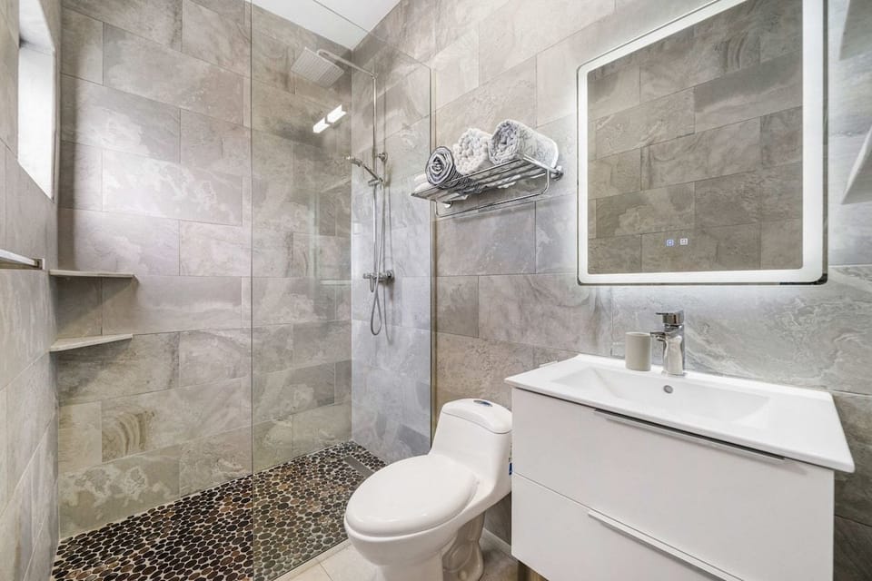 Complete spa-like bathroom featuring an LED backlit mirror, a contemporary white vanity, a toilet, and a glass-enclosed shower with a pebble floor.
