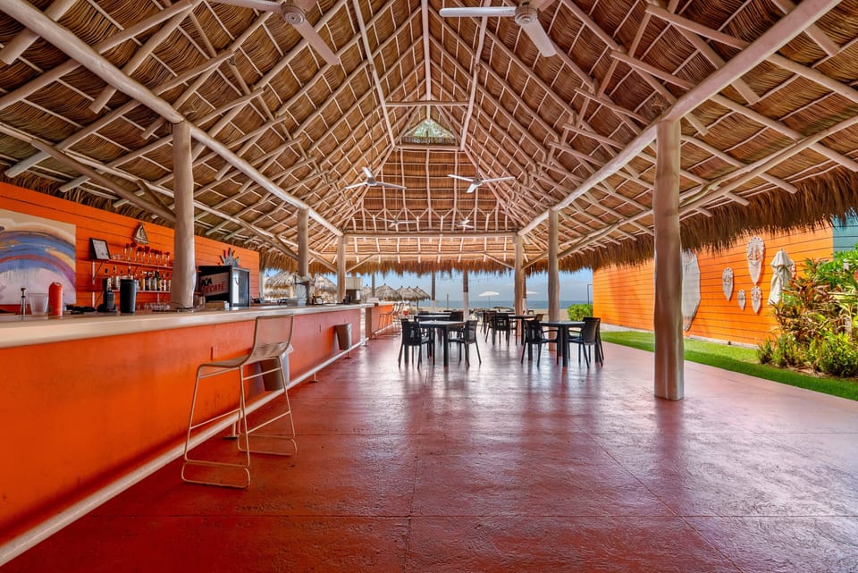 Spatious view under the soaring vaulted thatched palapa, highlighting the full-length orange bar counter, barstool seating, dining tables, and open breeze leading out toward the beach.