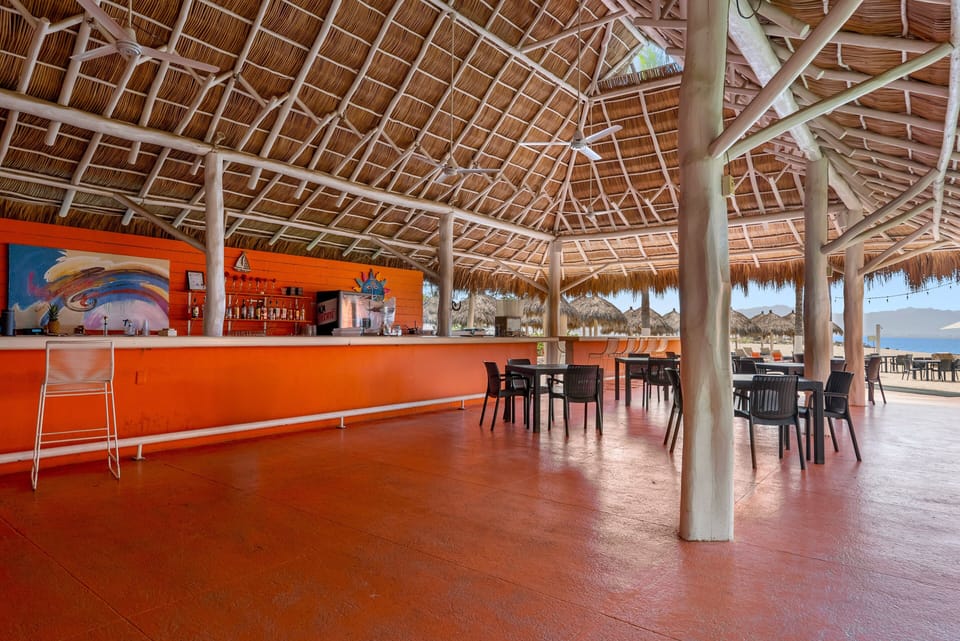 Vibrant orange beachfront bar and dining area under a massive high-ceiling thatched palapa, complete with ceiling fans, seating tables, and views looking out toward the ocean.